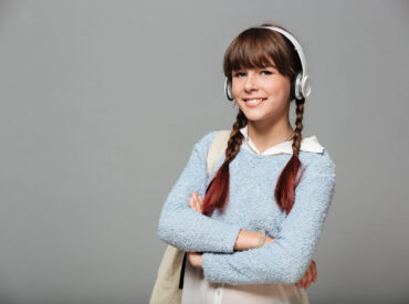 Portrait of a smiling young schoolgirl with backpack Portrait of a smiling young schoolgirl with backpack listening to music with headphones and looking at camera isolated over gray background